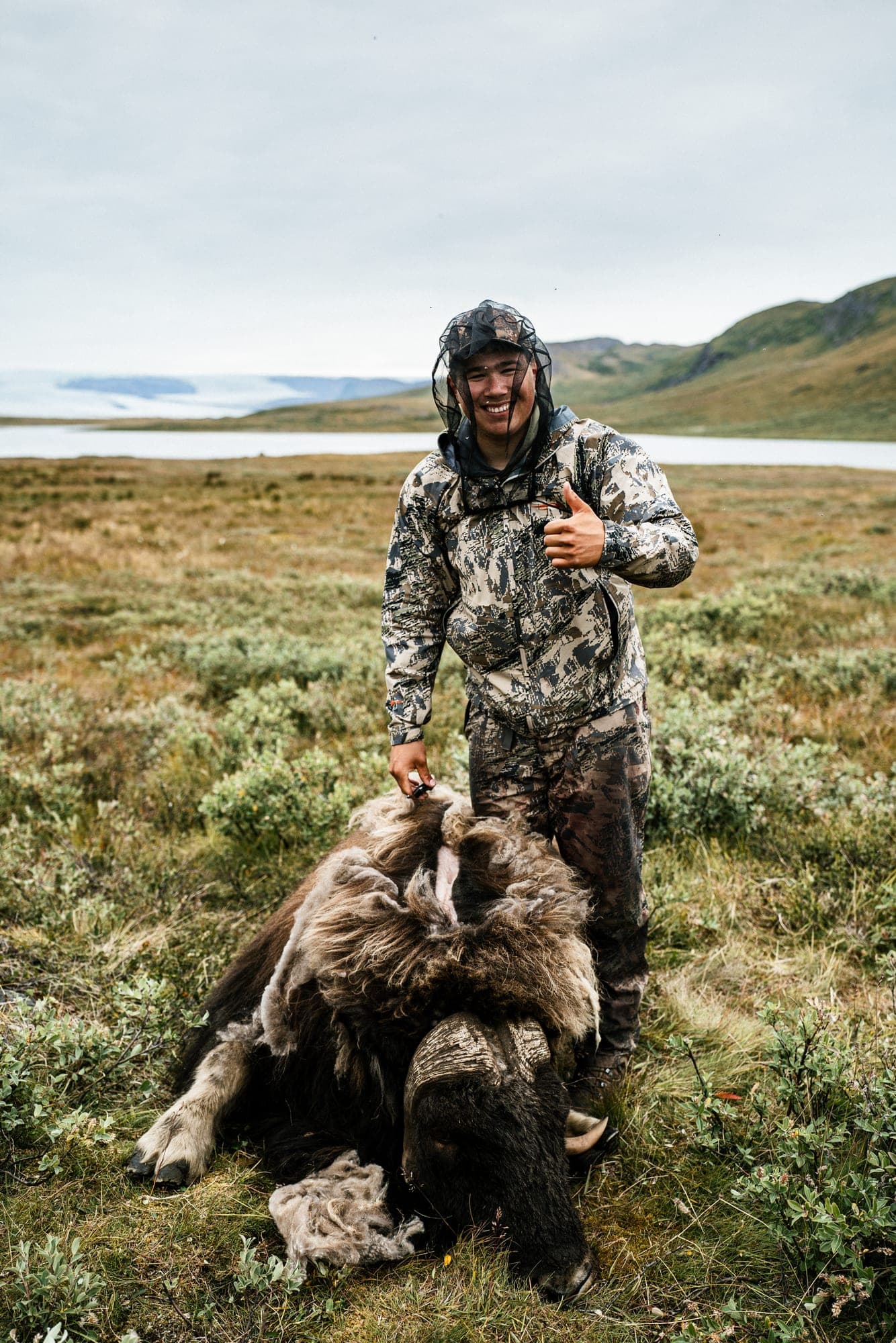 A smiling hunter in a mosquito net gives a thumbs up beside a musk ox