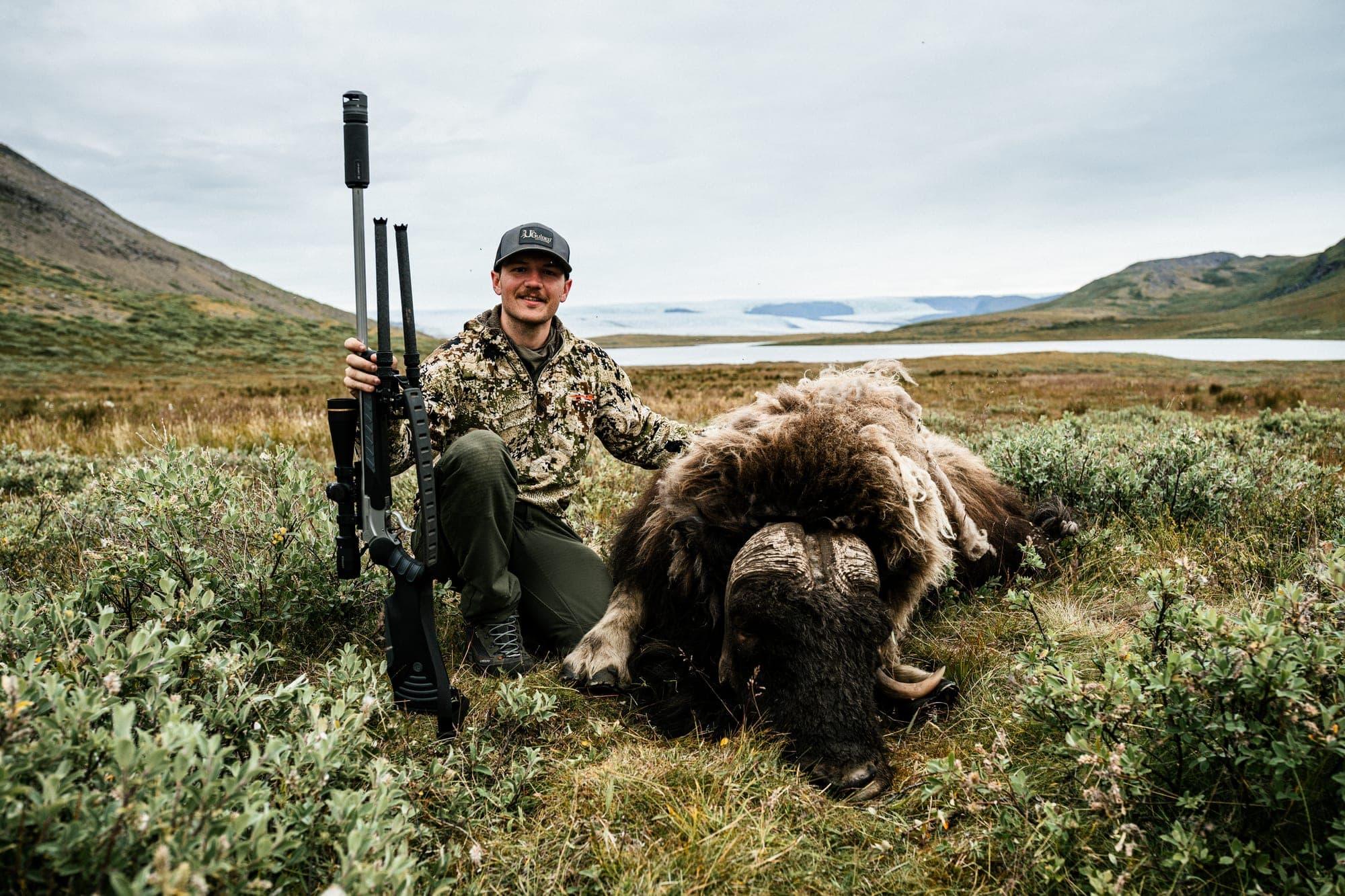 A rifle hunter kneeling beside a musk ox by a lake, the ice sheet behind