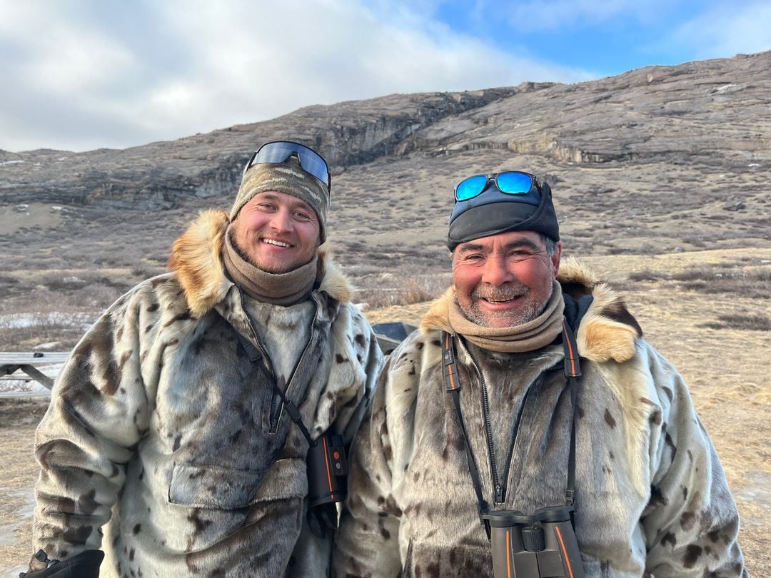 Two smiling men in spotted sealskin anoraks on the tundra, a rocky ridge behind them.