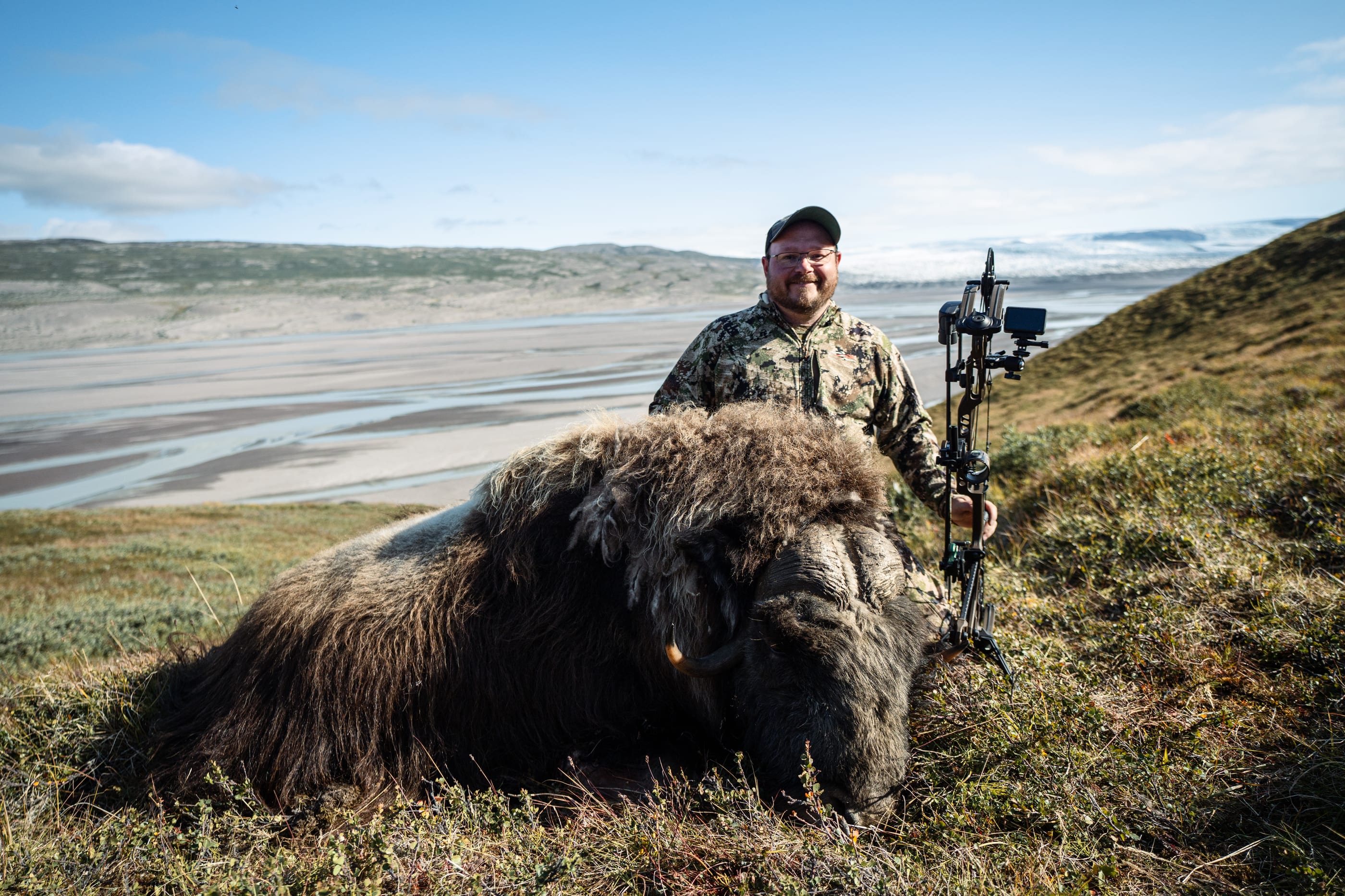 A bowhunter behind a musk ox on a slope, with the river delta and the ice sheet behind